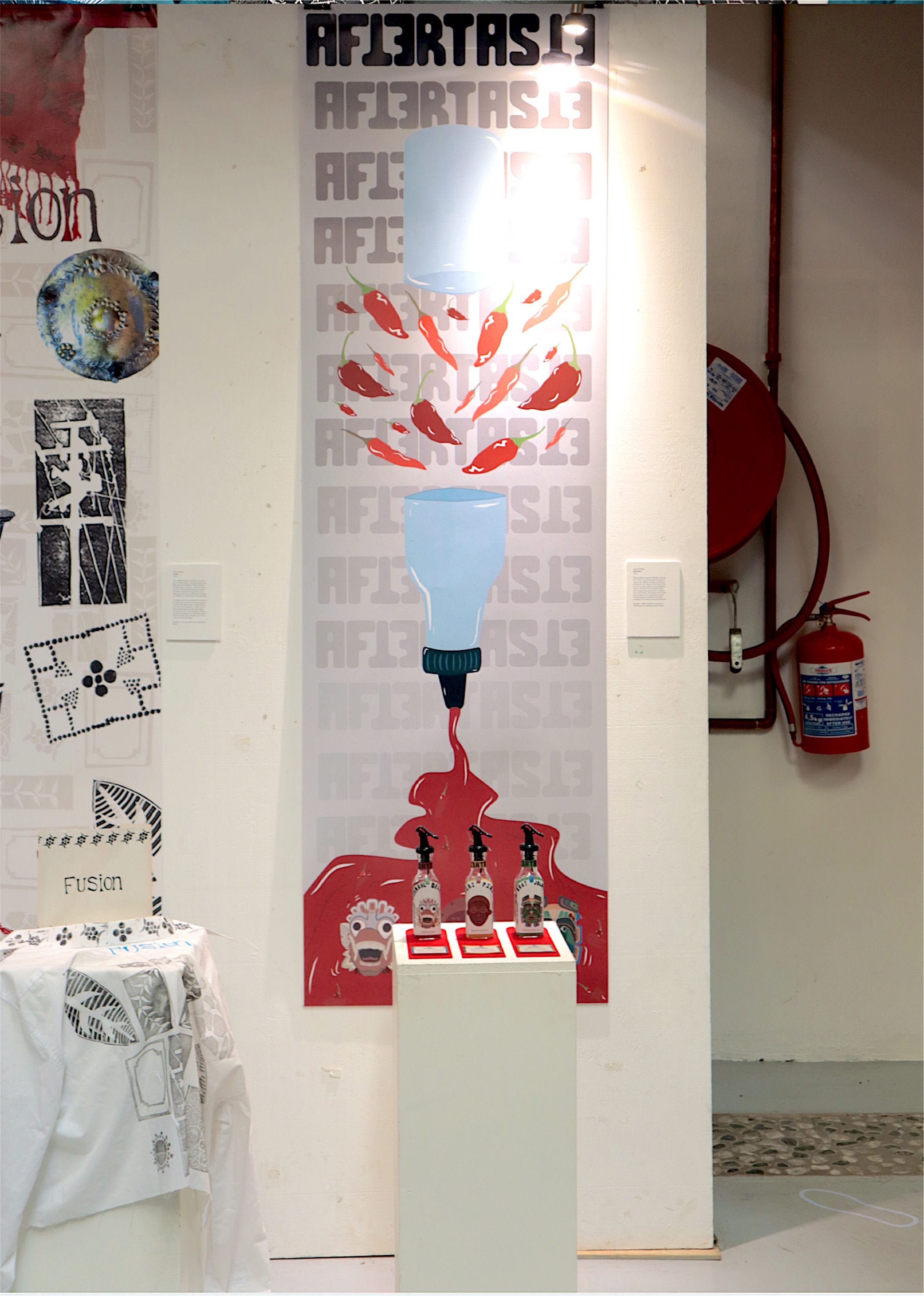 Exhibition detail: the Aftertaste wordmark wall with illustrated chillies and the hot sauce bottles on a plinth